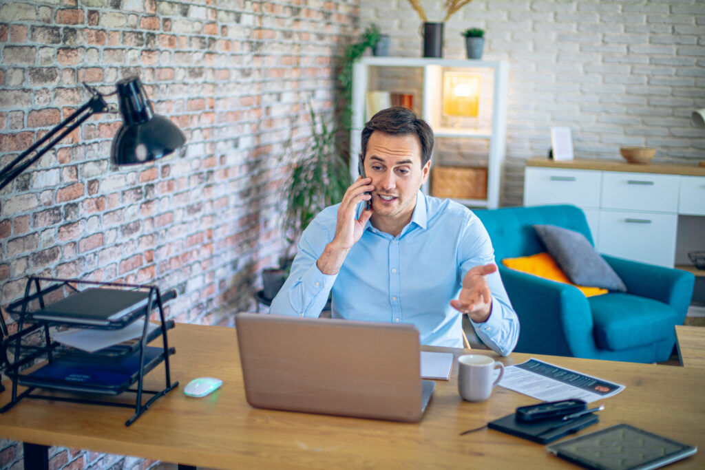 Young man talking on the phone in his home office in front of a laptop